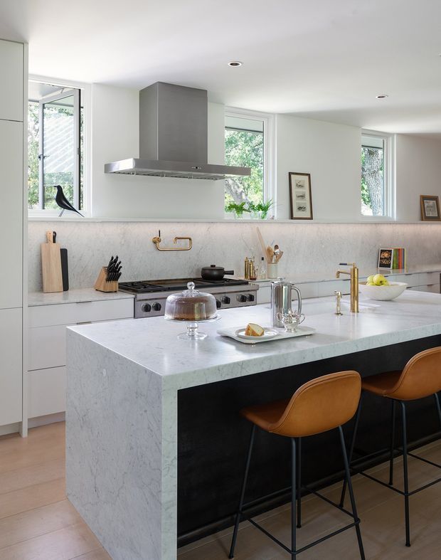 Bright contemporary kitchen featuring a large waterfall island with a pale marble-look benchtop and dark base, tan leather bar stools, white cabinetry, a freestanding stainless-steel rangehood, gas cooktop, integrated sink, and brass-toned tapware. Large windows bring in abundant natural light, while minimal framed artwork and greenery add subtle decoration.