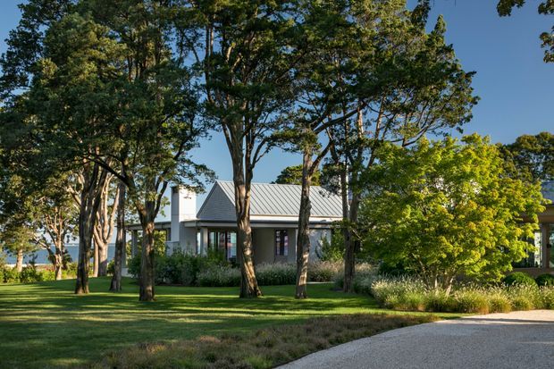 A low, contemporary house set within a mature landscaped garden. The building has a simple pale facade, a standing-seam metal gable roof, large glazed openings, and a broad lawn framed by established trees and ornamental planting. A curved pale driveway or path leads toward the house.