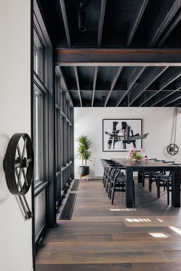 Industrial-style dining room with a long dark timber dining table and black chairs, framed by expansive black metal-framed glazing. The room features exposed black-painted ceiling beams and structure, dark metal detailing, a timber-look floor, white walls, a large abstract artwork, and a potted plant. A prominent wheel-and-belt mechanism is mounted on the wall, reinforcing the converted industrial character. Natural light creates strong rectangular patterns across the floor.
