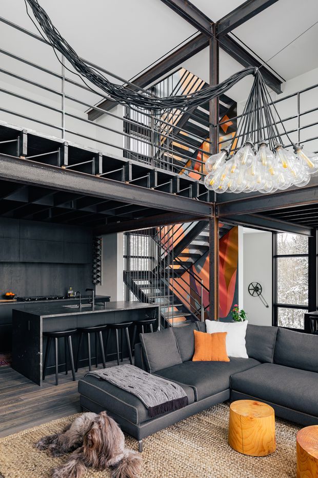 An open-plan loft-style interior combining a living room and kitchen beneath a dramatic double-height industrial structure. Exposed dark steel beams, an open steel stair with orange geometric wall accents, black-framed glazing, extensive visible cabling, and a clustered glass-bulb pendant create a bold warehouse-inspired character. The living area has a large dark sectional sofa, orange and white cushions, a textured rug, and timber stump side tables, while the compact dark kitchen includes an island with bar stools. A mezzanine level overlooks the space.