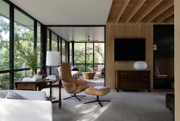 A refined mid-century-inspired living room with expansive black-framed glazing overlooking trees. The room features a grey carpet, pale upholstered seating, a tan leather lounge chair with matching ottoman, timber side furniture, and a built-in timber-panelled media wall with recessed shelving and a television. Exposed timber ceiling beams add warmth and architectural character.