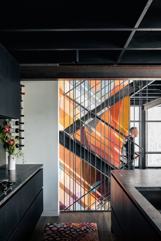 A dark, industrial-style interior looking toward a dramatic stairwell. Black fixed cabinetry and stone benchtops frame the foreground, with a wine bottle display and floral arrangement on the left. A black steel stair and balustrade rise through the centre against a vivid orange, white, and pale blue geometric feature wall. Exposed dark ceiling framing and a substantial steel beam reinforce the industrial character, while timber flooring and a patterned runner add warmth.