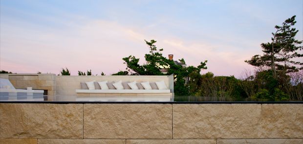 Exterior view of a landscaped rooftop or elevated terrace at dusk, featuring a long built-in white upholstered outdoor lounge against a pale stone privacy wall. The foreground has large textured beige stone cladding, with dense planting and trees behind the seating and a soft pink-blue sky beyond.