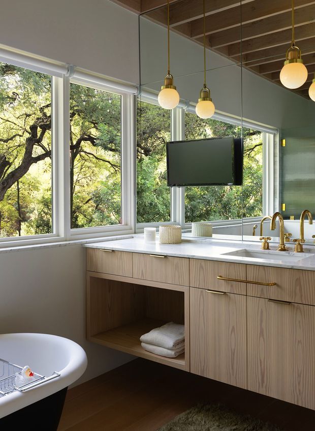 A refined contemporary bathroom with a freestanding bathtub, floating pale timber vanity, white stone benchtop, undermount basin, brass tapware, and a large mirrored wall. Floor-to-ceiling corner windows overlook leafy trees, while globe pendant lights hang from a warm timber-lined ceiling. A wall-mounted television and open towel storage are integrated into the vanity area.