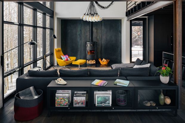 A dramatic, double-height living room with floor-to-ceiling black-framed glazing overlooking a wooded setting. The room features a dark sectional sofa, a black built-in media and storage console, a sculptural black fireplace wall with a compact wood-burning stove, a vibrant yellow lounge chair and ottoman, and an oversized glass-bulb chandelier. A mezzanine and dark timber structural elements are visible above the room.