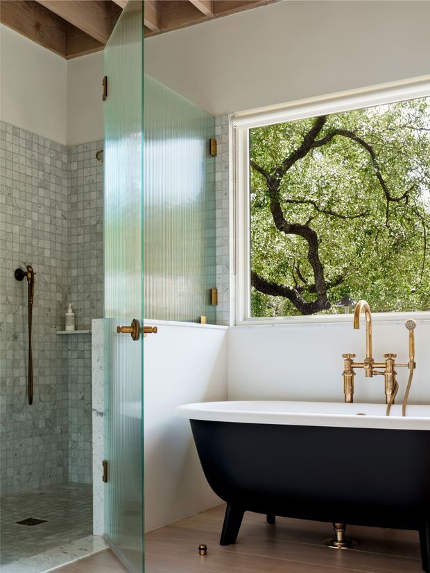 A refined bathroom featuring a black-and-white freestanding clawfoot bathtub beside a large window overlooking leafy trees. A walk-in shower is enclosed by vertically ribbed translucent glass, with pale square wall and floor tiles, warm brass plumbing and shower fixtures, white wall finishes, and a timber-lined ceiling detail.
