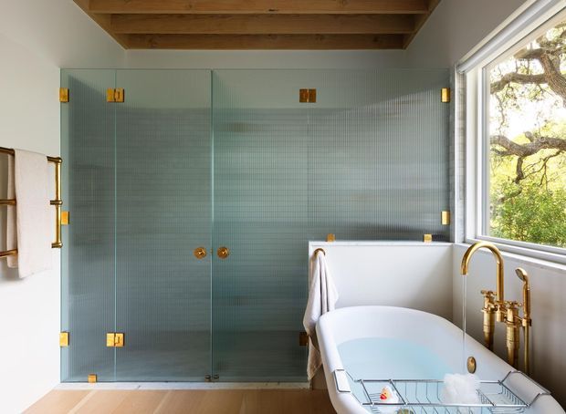 Bright, refined bathroom with a large frameless glass walk-in shower lined in pale blue-green textured tile, a freestanding white bathtub beside a large window, and a polished brass bath filler. Exposed timber ceiling beams, warm timber flooring, brass hardware, and abundant natural light create a warm contemporary aesthetic.