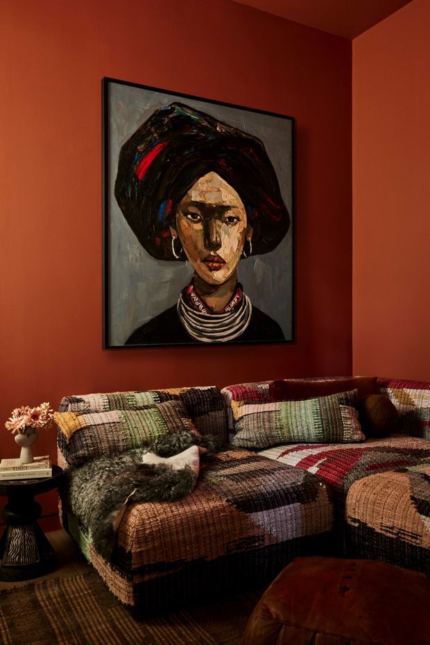A moody, richly coloured living room corner with deep terracotta-red walls, a large framed portrait artwork, and a low patchwork sectional sofa upholstered in multicoloured woven textiles. Layered cushions, a fur throw, a small side table with flowers and books, a patterned rug, and a brown leather ottoman create a warm, eclectic atmosphere.