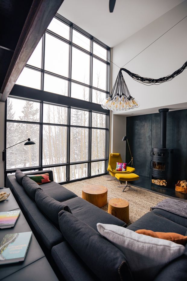 A dramatic double-height living room with expansive black-framed floor-to-ceiling glazing overlooking a wooded landscape. The room features a large dark charcoal sectional sofa, bright mustard-yellow lounge chair and ottoman, a black freestanding wood-burning stove set against a dark textured feature wall, round timber coffee tables, a patterned rug, and a sculptural chandelier with exposed black cords. The space has a modern, industrial character with abundant natural light and strong contrast.