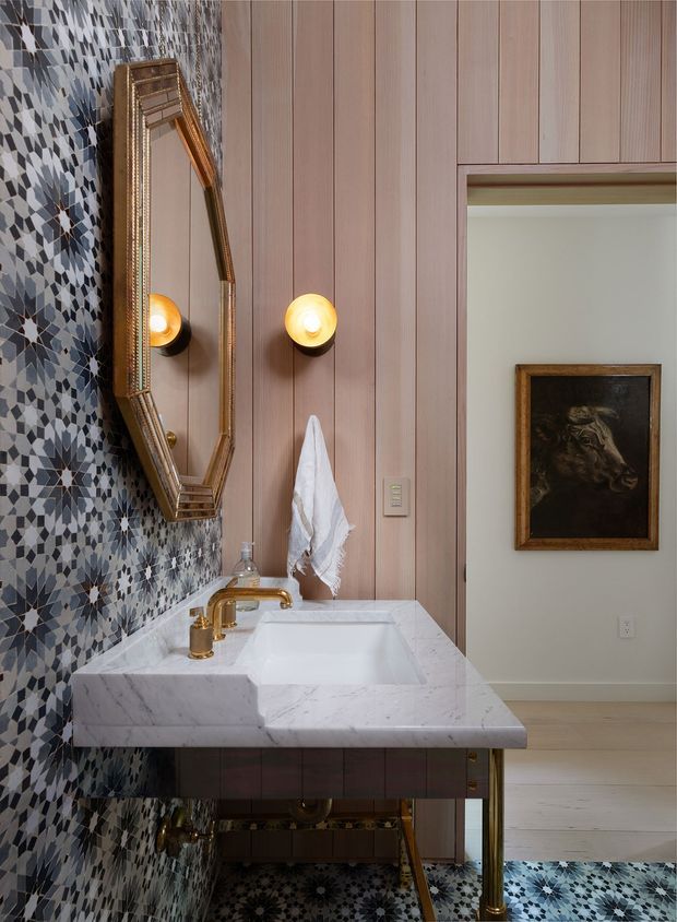 A richly detailed bathroom vanity area with a white marble countertop and integrated rectangular basin, polished brass tapware and legs, a large faceted vintage-style mirror, warm globe wall sconces, pink-toned vertical timber wall panelling, and intricate blue, grey, and white patterned tiles covering the feature wall and floor. A framed artwork is visible through the open doorway.