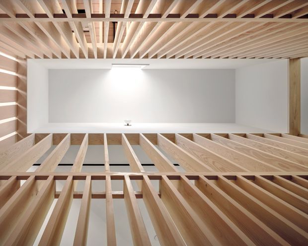 Upward-looking architectural view through a tall interior void, framed by a dense grid of exposed pale timber beams and rafters. White walls and a bright skylight or clerestory opening create a clean, luminous composition with strong linear perspective and visible structural framing.