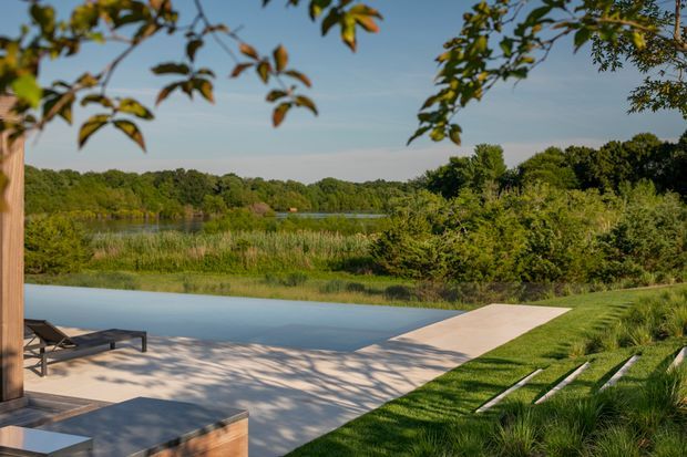 A landscaped outdoor pool area beside a natural pond or wetland, with a broad pale paved pool terrace, timber lounge furniture, a narrow walkway leading toward the water, and layered grasses and shrubs. Mature trees frame the view, creating a secluded, natural setting.