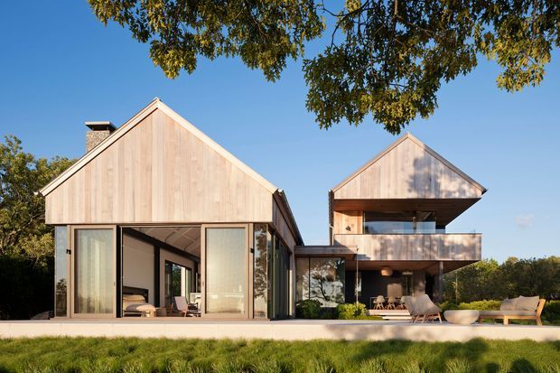 Exterior view of a contemporary multi-volume residence with two steep gabled forms clad in light timber, extensive floor-to-ceiling glazing, and a cantilevered upper-level balcony. The home opens onto a landscaped lawn with outdoor seating and mature trees surrounding the site.