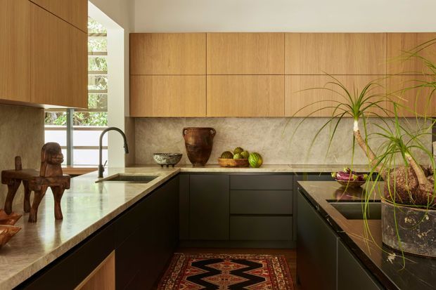 Warm, refined contemporary kitchen with an L-shaped island and peninsula arrangement, extensive vertical-grain timber cabinetry, dark lower cabinets, and a mottled stone backsplash and benchtop. A tall window brings in natural light, while a sculptural timber stool, pottery, fruit, rug, and large indoor plants add character. The kitchen has a calm, earthy, mid-century-influenced atmosphere.
