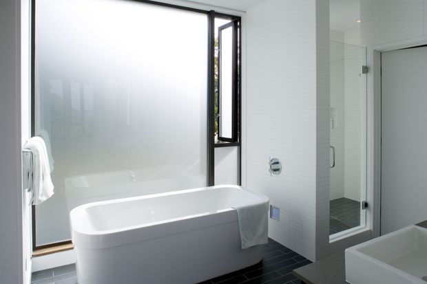 Bright contemporary bathroom featuring a freestanding white bathtub beside a large frosted-glass window, white tiled walls, dark floor tiles, and a glazed shower enclosure.
