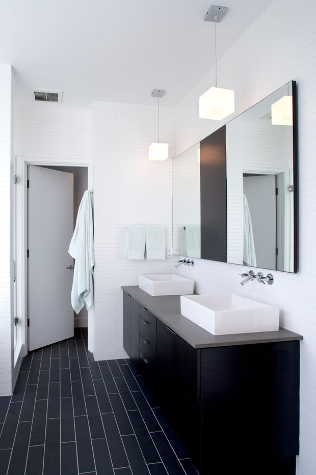 Bright contemporary double-vanity bathroom with white horizontal wall panelling, dark tiled flooring, a long dark timber vanity, two white vessel basins, chrome tapware, and a large framed mirror. Two geometric pendant lights hang above the vanity. An open doorway and towels are visible at the rear.