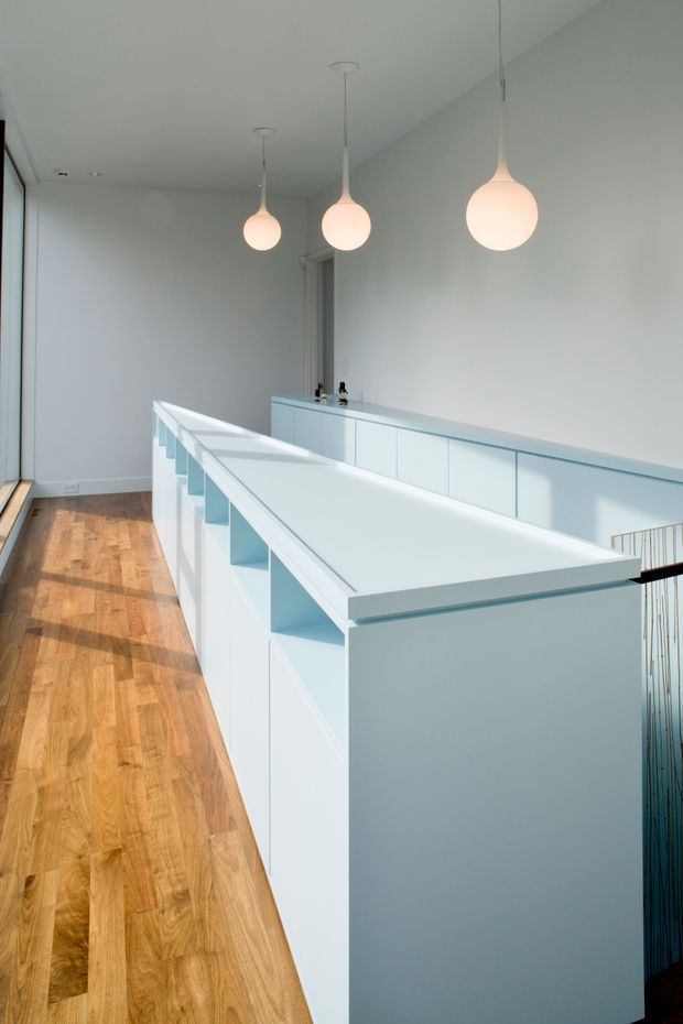 A bright minimalist interior hallway or upper-level landing with extensive white built-in cabinetry and balustrade-style joinery. Three softly glowing pendant lights hang from the white ceiling, while warm timber flooring contrasts with the cool white surfaces. A stair opening with a dark vertical balustrade detail is visible at the right.