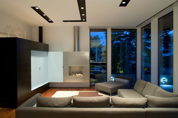 Contemporary living room with a large sectional sofa, built-in dark timber cabinetry, pale wall-mounted joinery, and a minimalist linear fireplace. Floor-to-ceiling glazing wraps the corner, while recessed linear ceiling lights and warm timber flooring create a refined evening interior.