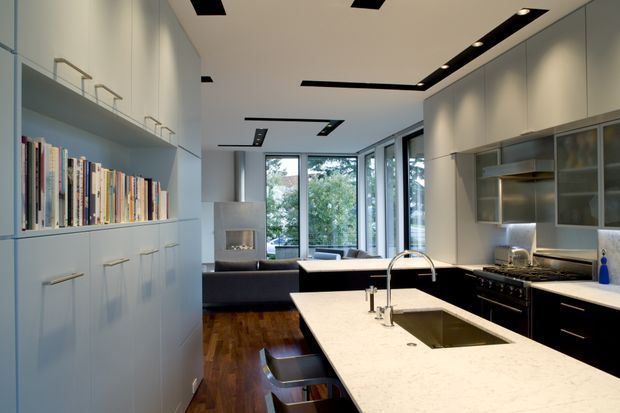 Contemporary open-plan kitchen with a large pale stone island and integrated sink, dark lower cabinetry, stainless-steel appliances, and extensive built-in white cabinetry with open shelving. The kitchen opens directly to a living area with a fireplace and large glazed doors overlooking trees. Recessed linear black ceiling lighting and warm timber flooring add contrast.