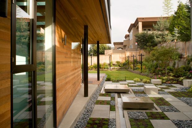 A contemporary residential courtyard viewed from beneath a deep timber-clad overhang. The building features warm horizontal wood wall cladding, dark slender posts, expansive glazing, and a paved garden path composed of large square concrete or stone stepping slabs set among grey gravel, planted groundcover, and structured garden beds. The courtyard is enclosed by timber and masonry fencing with neighbouring homes visible beyond.