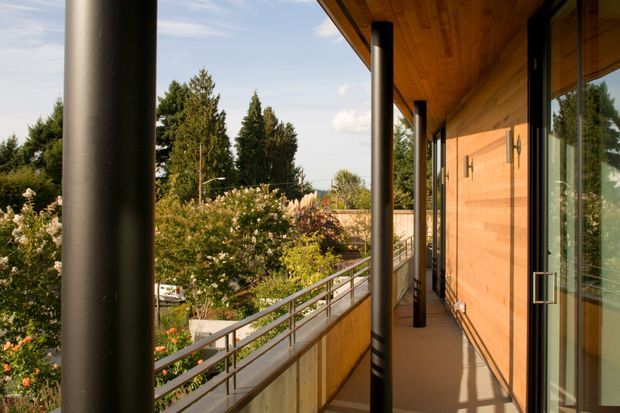 A covered exterior balcony or terrace runs along a timber-clad building, with a warm timber soffit and wall, dark cylindrical structural columns, metal balustrade, and large glazed doors. The elevated walkway overlooks landscaped suburban gardens and trees.