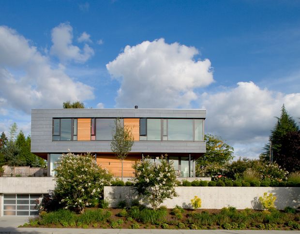 A modern two-storey residence with a long rectangular form, flat roof, extensive glazing, grey horizontal exterior cladding, and warm timber wall panels. The house sits above a landscaped front garden with a concrete retaining wall and an integrated garage.