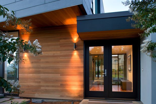 Contemporary residential entrance viewed from outside, featuring a warm horizontal timber-clad wall and soffit, dark-framed glazed double doors, crisp geometric roof forms, and integrated exterior lighting. Landscaping partially frames the entry, while the interior hallway is visible through the glazing.