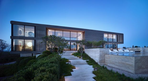 Contemporary two-storey residence photographed at dusk, featuring a long rectilinear form, dark horizontal exterior cladding, extensive floor-to-ceiling glazing, and a recessed central entry. A landscaped garden and broad concrete path lead toward the house, while a raised stone-clad terrace or pool area extends at the right.