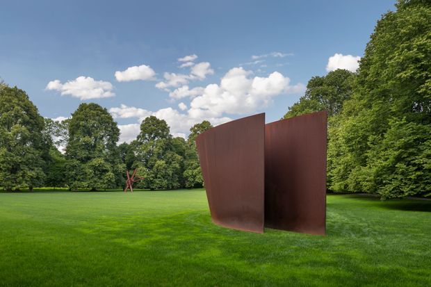 An outdoor sculpture installation consisting of two tall, curved, rust-coloured steel plates standing in a broad manicured lawn, surrounded by mature leafy trees under a partly cloudy blue sky. A smaller red abstract sculpture is visible in the distant landscape. The image is a landscape-oriented exterior view with the sculpture as the focal point.