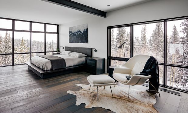 A contemporary bedroom with expansive floor-to-ceiling black-framed glazing overlooking a snowy forest. The room features a low platform bed with dark bedding, a sculptural cream lounge chair and ottoman, a dark side table, timber flooring, a black exposed ceiling beam, and restrained artwork. The monochrome palette and extensive glazing create a calm, refined retreat.