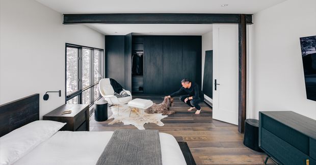 A modern bedroom viewed from the sleeping area, with a low bed, dark timber flooring, white walls, large windows, dark built-in cabinetry, and exposed steel beams. A small seating area with an armchair and ottoman occupies the far end, while a dark sideboard and wall-mounted television are visible on the right.