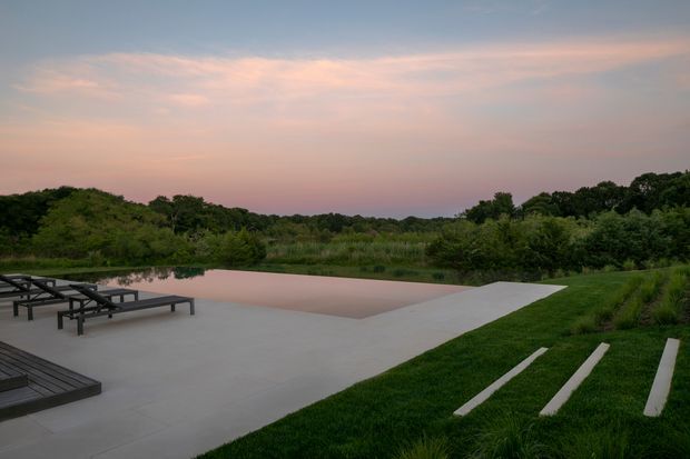 A tranquil landscaped outdoor setting at dusk with a minimalist rectangular reflecting pool or swimming pool, broad pale concrete paving, dark timber decking and lounge chairs, and linear stepping stones set into a manicured lawn. Dense vegetation forms a natural backdrop beneath a soft pink and blue evening sky.