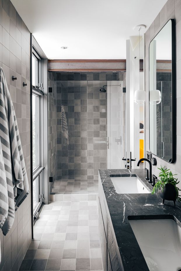 A narrow, bright contemporary bathroom with a walk-in tiled shower at the rear, a long dark stone vanity with two integrated white basins, black curved tapware, a large wall mirror, and a tall window along the left side. Grey tiled walls and floor are contrasted by exposed timber ceiling beams and black-framed glazing.
