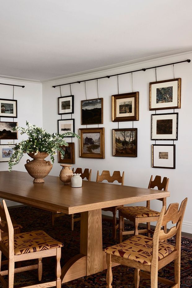 A traditional dining room with a large rectangular timber dining table surrounded by carved timber chairs with patterned upholstered seats. The white walls are decorated with an extensive gallery of framed artwork hung from black picture rails. A large woven vase with leafy branches and a smaller decorative vase sit on the table, while a patterned area rug covers the floor. The room has white walls, ornate ceiling cornices, and warm natural timber furnishings.