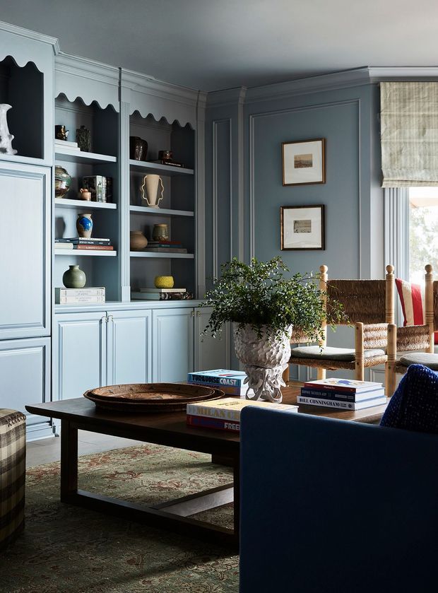 A refined, traditional living room with built-in pale blue-grey cabinetry and open shelving featuring decorative scalloped detailing. The room has paneled walls, a dark wood rectangular coffee table, upholstered seating, woven dining-style chairs, framed artwork, a patterned rug, and a large ornate planter. Natural light enters through a window with a textured Roman blind, creating a calm, sophisticated atmosphere.