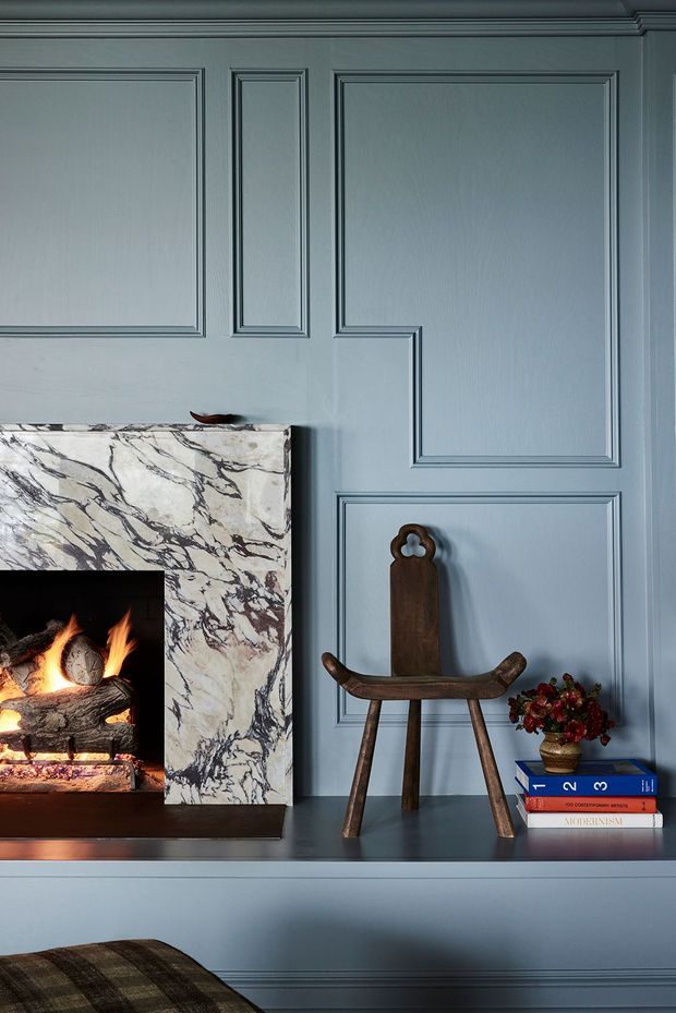 A moody, refined interior detail featuring a blue-grey panelled wall and built-in mantel. A fire burns in a fireplace framed with heavily veined white marble, while a small rustic wooden chair, stacked books, and a floral arrangement sit on the dark mantel surface. Decorative mouldings and a deep blue-grey palette create a classic, intimate atmosphere.