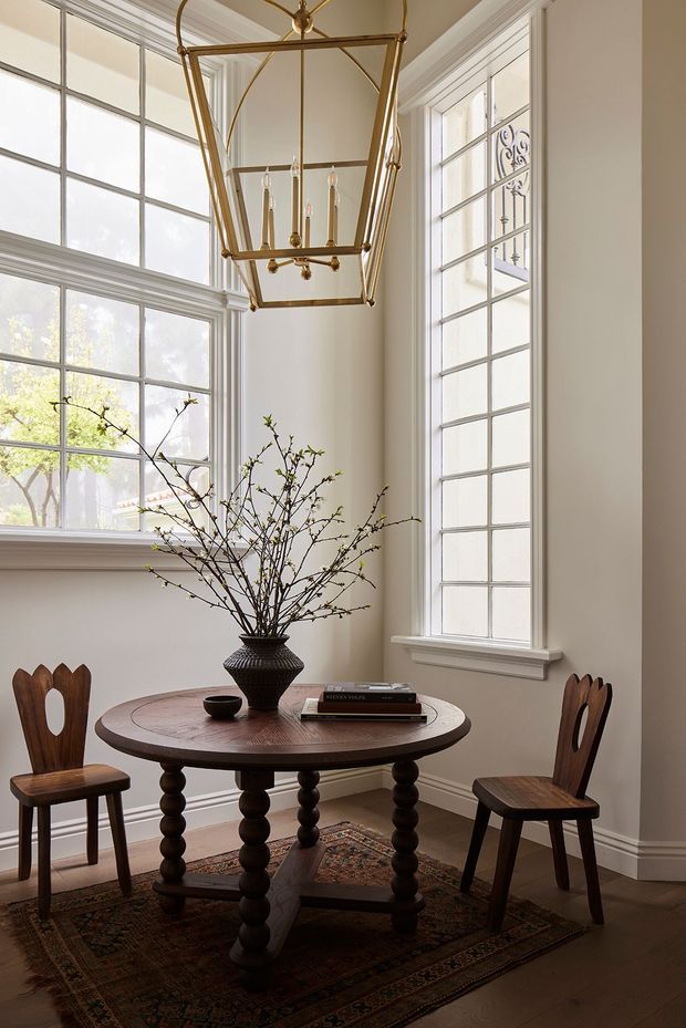A refined dining nook with a round dark timber pedestal table, two carved timber chairs, a decorative vase with branches, and a patterned rug. Tall multi-pane windows with substantial white painted trim fill the corner with natural light, while an oversized brass lantern pendant adds a traditional architectural accent.