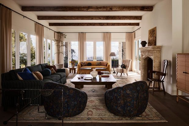 A refined, generously proportioned living room with tall windows, full-length curtains, exposed dark timber ceiling beams, and a carved stone fireplace. The room is furnished with a dark striped sofa, patterned lounge chairs, a central wooden coffee table, a mustard-toned sofa, and a vintage-style rug. Abundant natural light and layered traditional furnishings create a warm, elegant atmosphere.