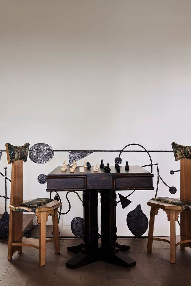 A compact dining area featuring a dark wood pedestal dining table with two carved wooden chairs upholstered in patterned fabric. Sculptural black and metallic decorative objects are arranged on the tabletop and displayed against a pale wall, with dark timber flooring and a restrained, gallery-like composition.