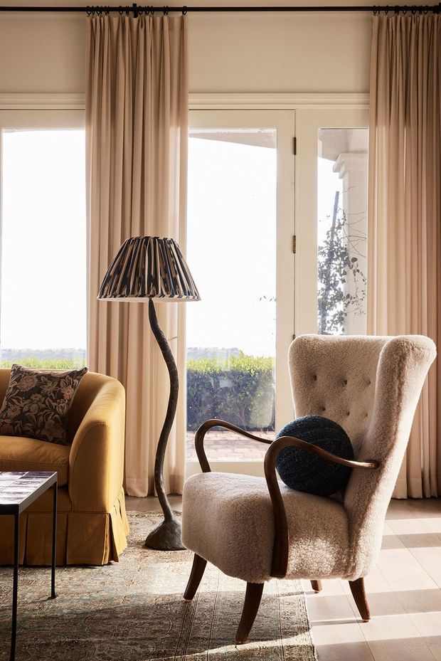 A warmly styled sitting room with tall glazed doors and windows dressed in full-length beige curtains. The room features a mustard upholstered sofa with a patterned cushion, a sculptural floor lamp, a plush high-back armchair, a dark round cushion, a patterned area rug, and pale tiled flooring. Abundant daylight and traditional wall mouldings create a refined, cozy atmosphere.