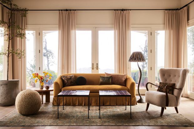 Warm, refined living room with a mustard velvet sofa, sculptural armchair, paired black-framed coffee tables, textured rug, tall curtains, and large multi-panel windows providing abundant natural light. Indoor plants and layered furnishings create an inviting, softly eclectic atmosphere.