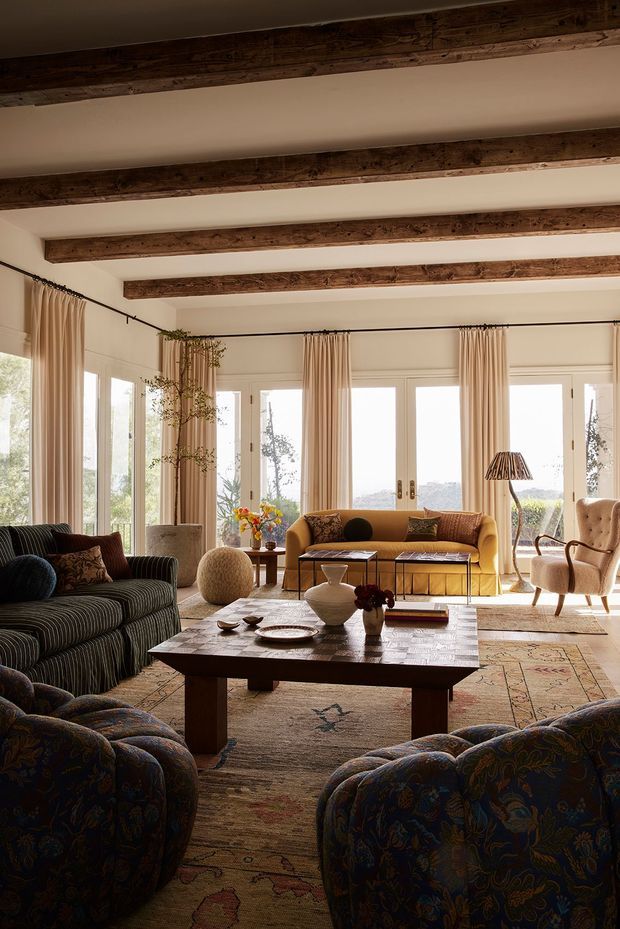 A warm, richly layered living room with exposed dark timber ceiling beams, cream walls, tall glazed doors and windows dressed with full-height beige curtains. The room is furnished with patterned upholstered sofas and armchairs, a mustard-toned sofa, a substantial dark timber coffee table, vintage-style rugs, and decorative lamps and accessories. The composition feels relaxed, refined, and traditionally inspired, with abundant natural light from the garden-facing glazing.