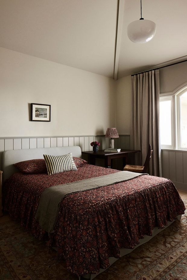 A warmly styled bedroom with a large bed dressed in a dark red and brown patterned quilt, striped and patterned cushions, and a muted green runner. The room features pale walls, grey-painted vertical wall panelling, a simple white upholstered headboard, dark wooden bedside furniture, heavy taupe curtains, an arched window, a patterned area rug, and a white pendant light. The overall atmosphere is intimate, traditional, and softly lit.
