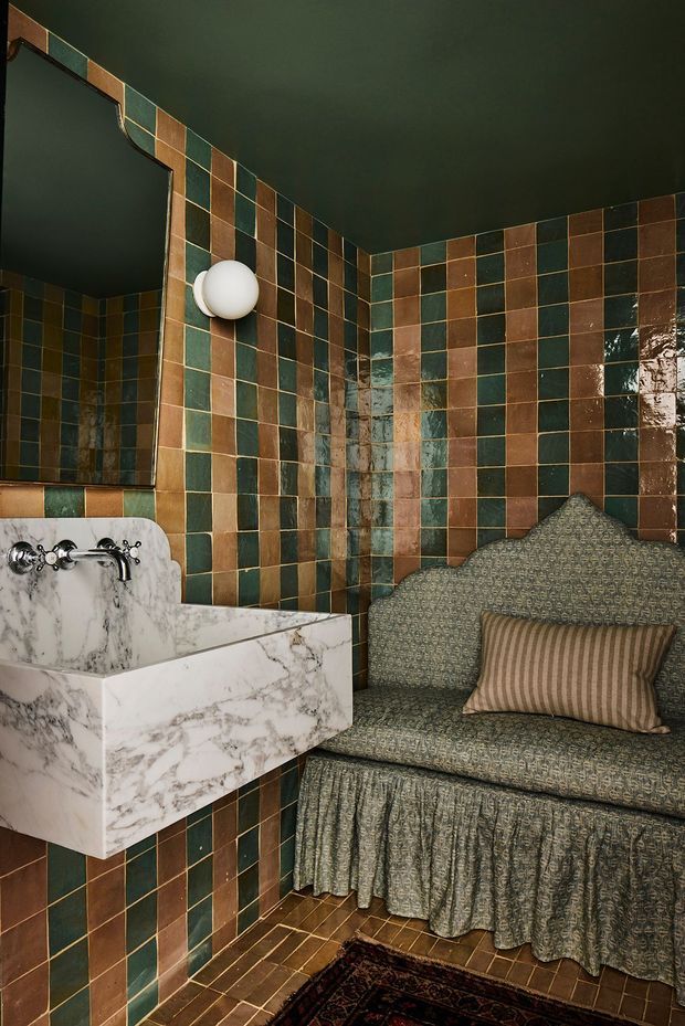 A compact, richly styled bathroom featuring glossy square wall and floor tiles in alternating deep green, brown, and muted terracotta tones. A sculptural marble wall-mounted basin with exposed chrome plumbing sits beneath a large mirror and spherical wall light. A patterned upholstered bench with a scalloped back and gathered skirt occupies the corner, accompanied by a striped cushion. The room has a dark ceiling, ornate traditional detailing, and a moody, eclectic atmosphere.