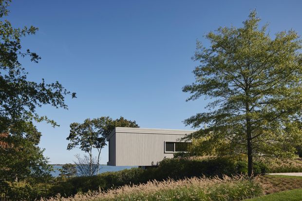 A minimalist rectangular building viewed from a landscaped exterior setting. The structure has a simple low-pitched or flat roof, pale vertically ribbed cladding, and a dark horizontal window. Mature trees, ornamental grasses, shrubs, and a glimpse of blue water or distant landscape surround the building under a clear sky.