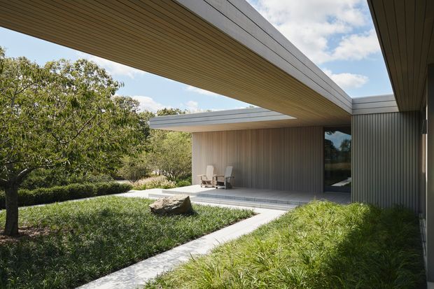 A low-profile contemporary house arranged around a landscaped courtyard. Broad cantilevered roof planes and soffits frame an outdoor seating area, while vertically ribbed wall cladding and large dark glazed openings define the facade. Pale paved paths pass through ornamental grasses and clipped planting, with mature trees beyond.