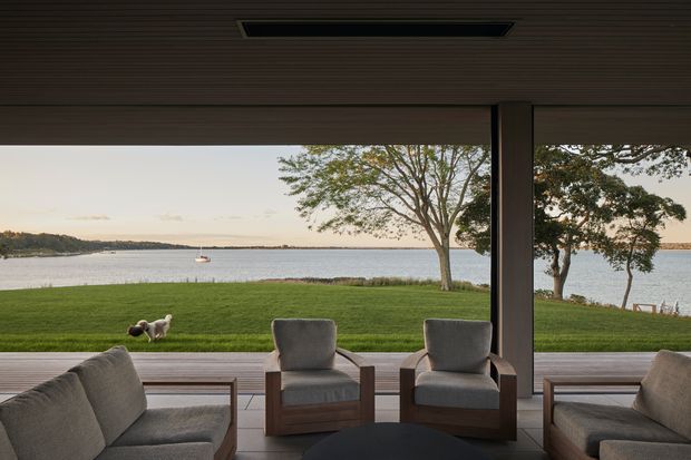 A covered waterfront outdoor living area framed by broad timber-clad roof soffits and substantial structural posts. The space contains cushioned lounge chairs and sofas arranged around a low dark coffee table, opening directly onto a manicured lawn and calm water beyond. A rectangular recessed ceiling opening adds a subtle architectural feature. The setting is relaxed, refined, and strongly connected to the waterfront landscape.
