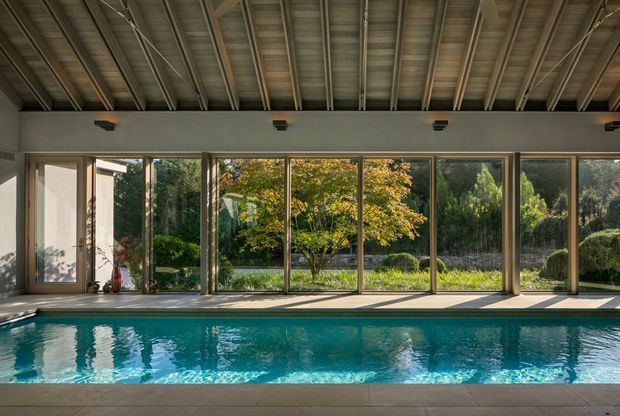 A covered indoor swimming pool enclosed by extensive floor-to-ceiling glazing, with a long rectangular pool, tiled pool surround, exposed metal roof structure, and views to a landscaped garden. The space is bright and strongly connected to the outdoors.