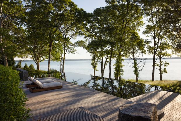 A spacious timber deck overlooking calm water, surrounded by mature trees and natural vegetation. The deck includes low-profile outdoor lounge chairs and a rustic stump-like side table, creating a serene waterfront outdoor retreat.