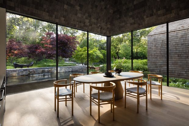 A refined dining room with a large round wooden dining table and matching timber chairs, enclosed by expansive floor-to-ceiling glazing. The room opens visually onto a landscaped garden with a stone-edged water feature, while dark textured timber cladding wraps the ceiling and an adjacent wall. Warm sunlight, pale flooring, and carefully integrated furnishings create a calm, nature-connected interior.